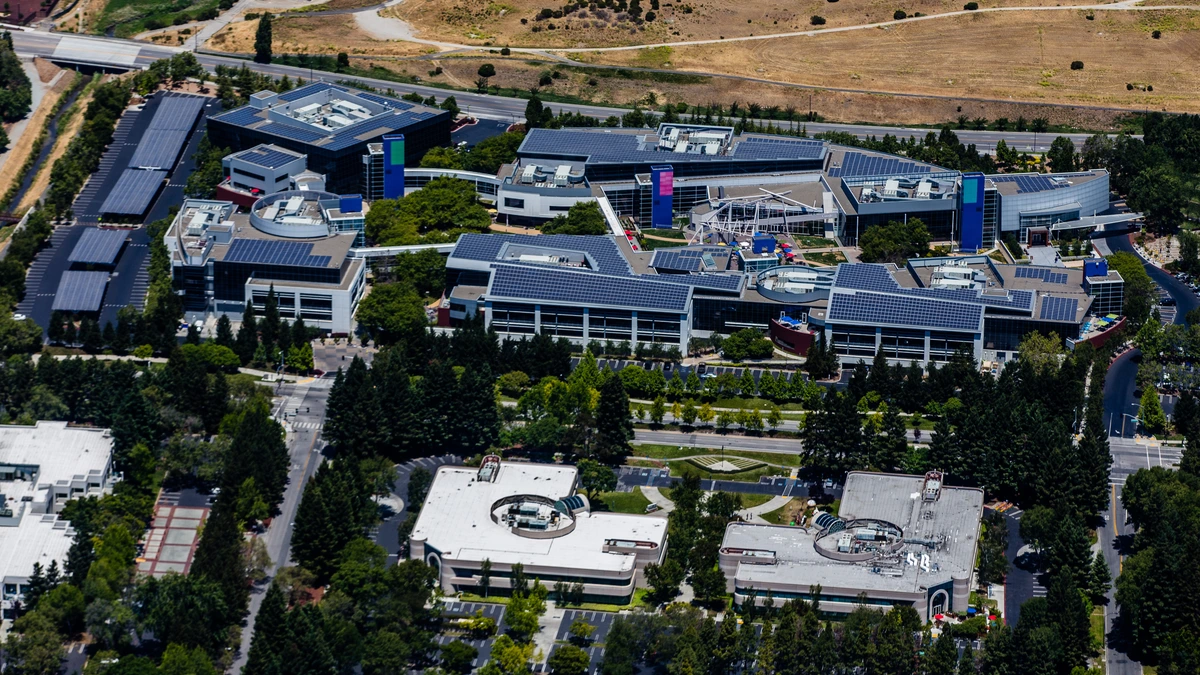 Aerial view of the Google campus in Mountain View, California, with low office buildings, solar panels on the roofs, tree-lined paths and dry hills behind