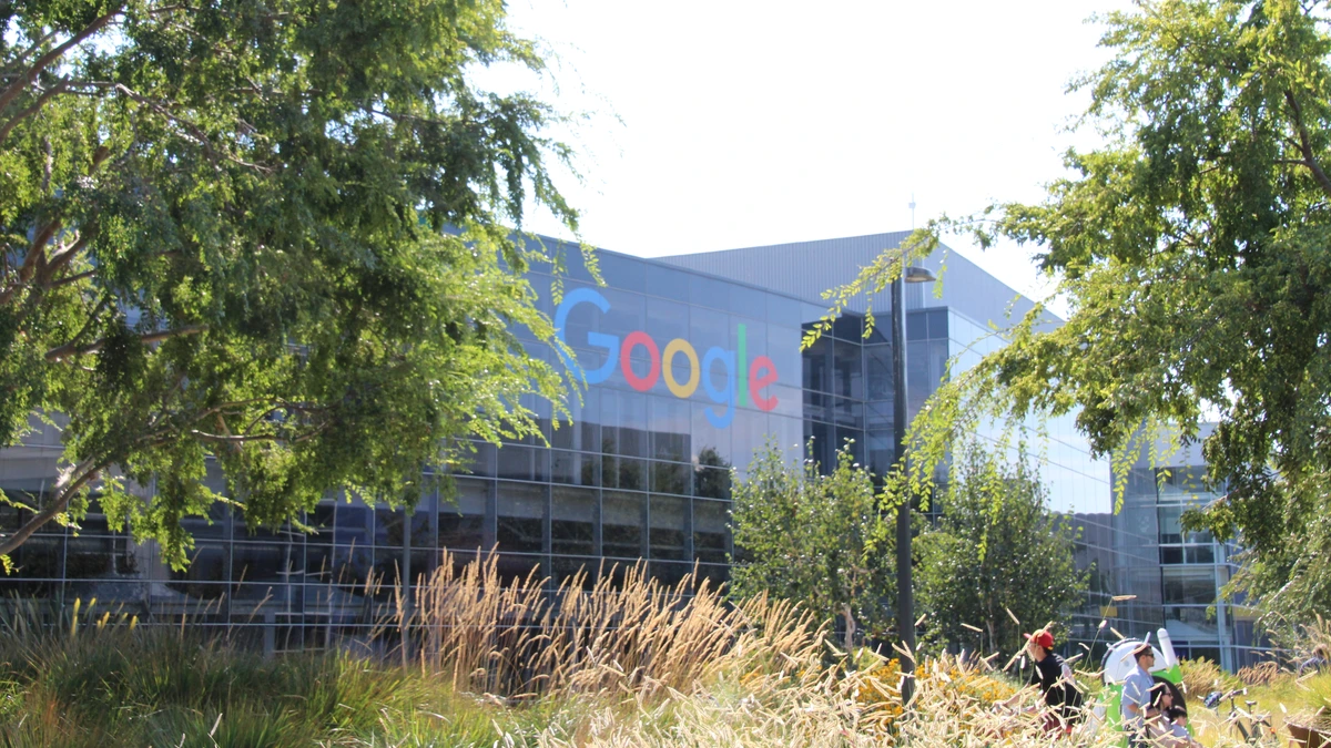 The glass-fronted Googleplex building in Mountain View seen through trees and tall dry grass, with the Google logo on the facade