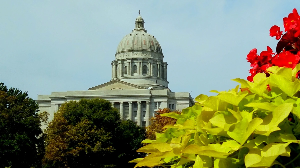 Domed Missouri State Capitol in Jefferson City seen over green trees, with red begonias and lime-green leaves in the foreground