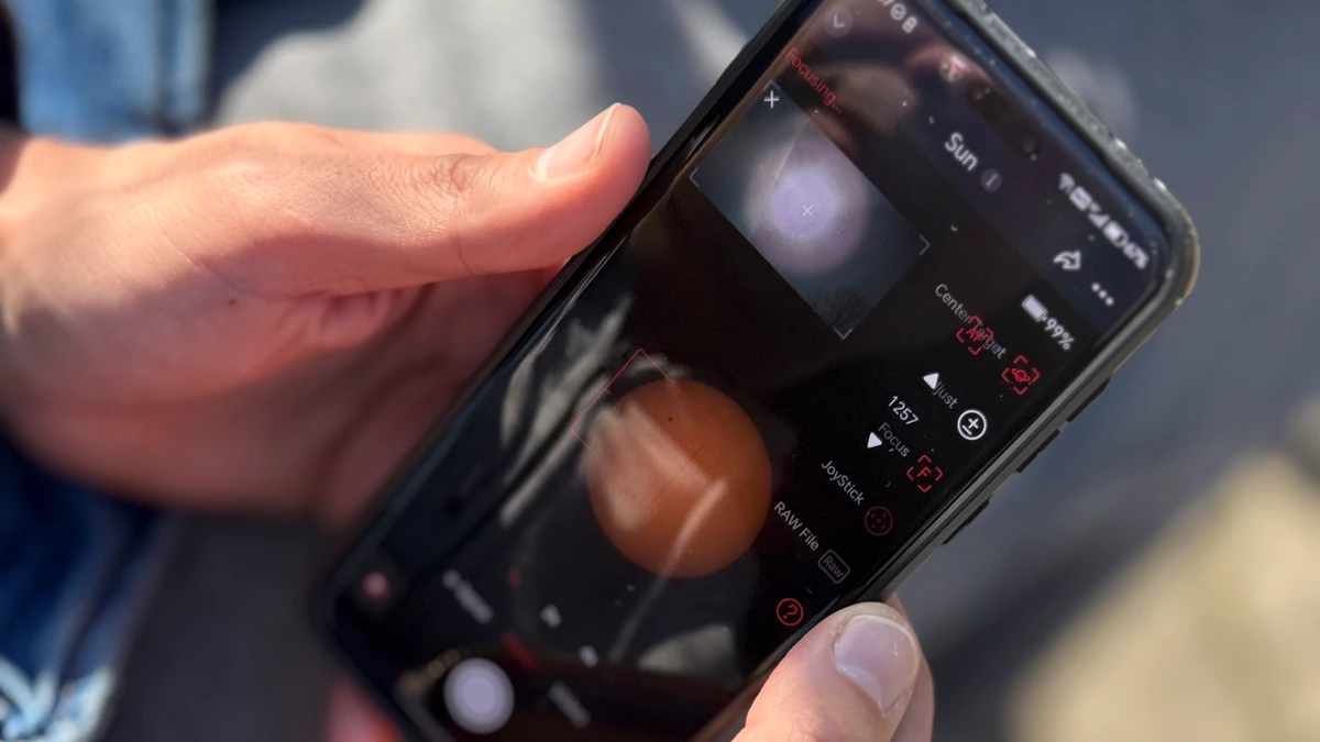 Two hands hold a smartphone showing a camera app with focus and exposure controls, over a denim jacket in bright sun