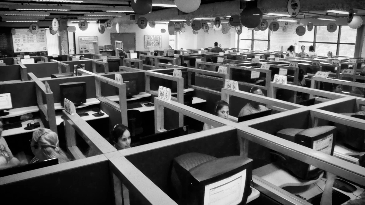 A black and white photograph looking across a large call centre floor of low partitioned booths, each with a boxy monitor, with several agents wearing headsets at their desks
