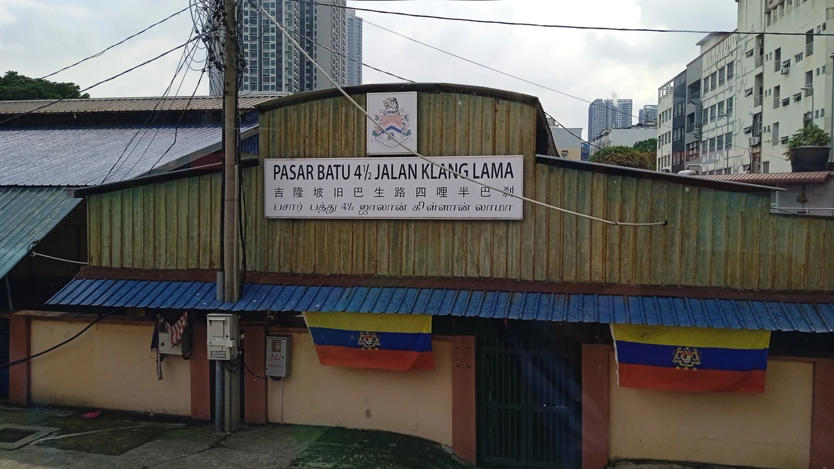 The trilingual entrance sign of Pasar Batu 4 1/2 on Jalan Klang Lama in Kuala Lumpur, reading the market's name in Malay, Chinese and Tamil above a shuttered market building with Malaysian flags hanging below