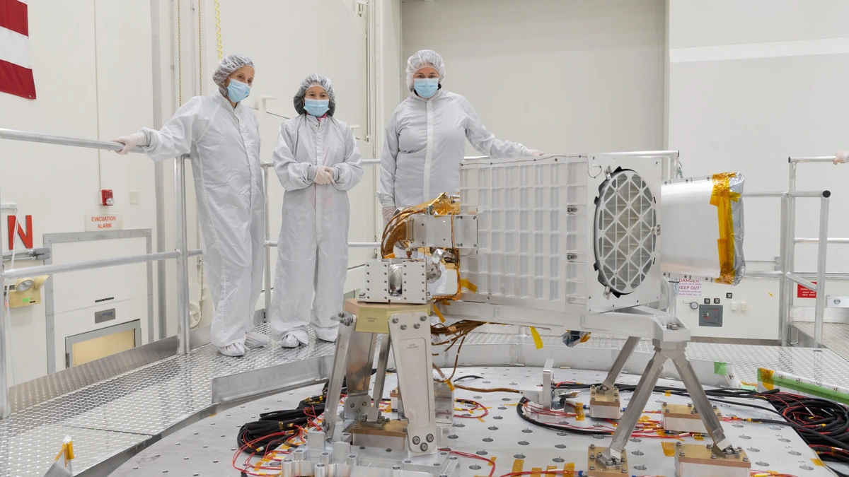 Three mission staff in white cleanroom suits standing beside the EMIT science instrument mounted on a vibration test table at NASA's Jet Propulsion Laboratory
