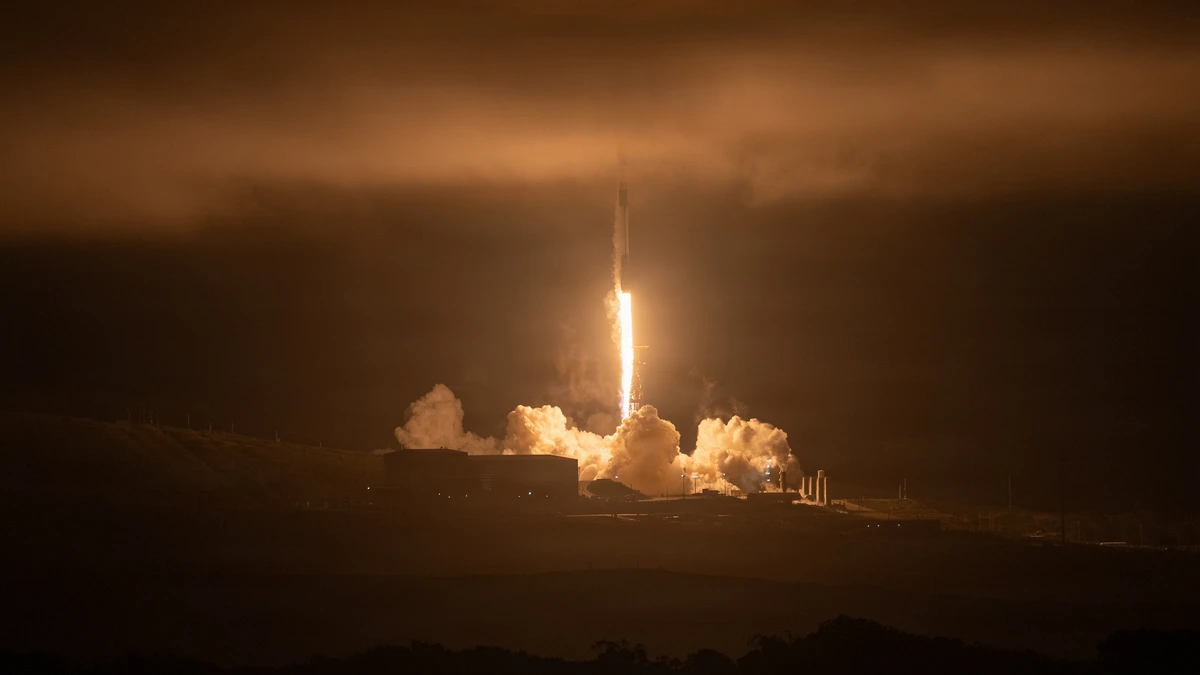 A SpaceX Falcon 9 rocket lifts off at night from a launch pad, its exhaust glowing orange against low cloud