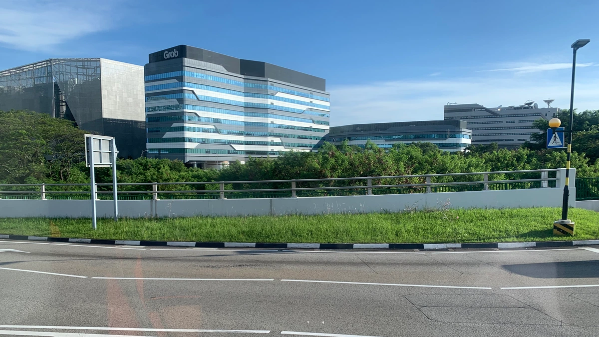 Grab's headquarters at one-north in Singapore, a striped blue and white office block with a Grab sign on the roof, seen across a road, a railing and a grass verge