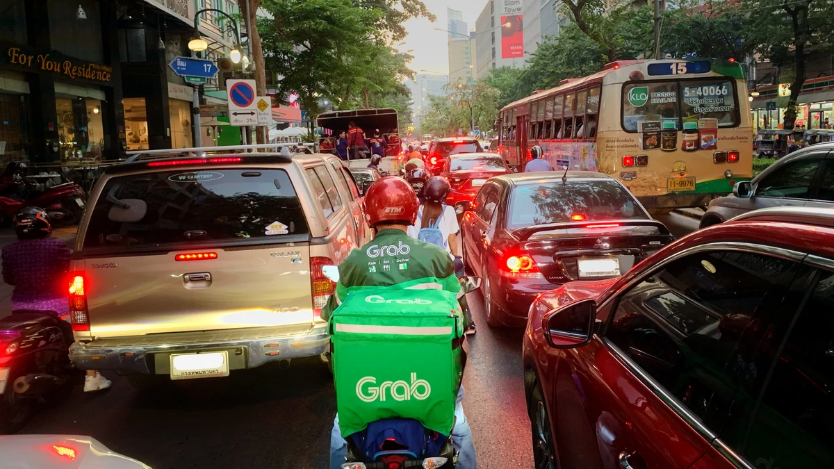 A Grab delivery rider in a green Grab jacket and red helmet, with a green Grab delivery box on the motorcycle, waiting in heavy evening traffic behind cars and a city bus in Bangkok