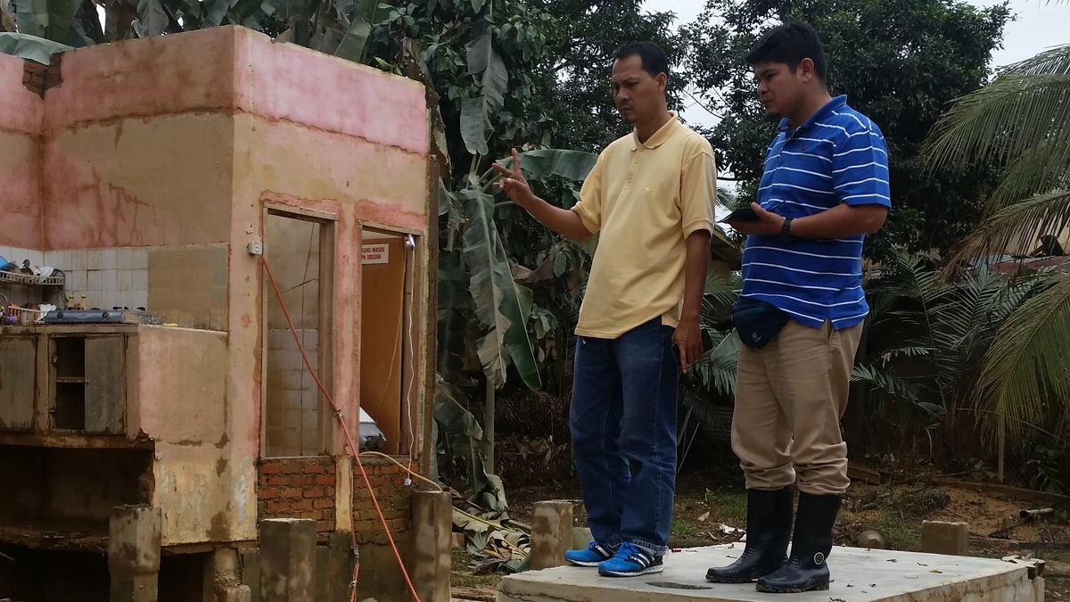Two men stand on the concrete steps of a flood-wrecked building in Gua Musang, Kelantan, one pointing at the damaged pink walls while the other holds a phone, with mud and debris on the ground
