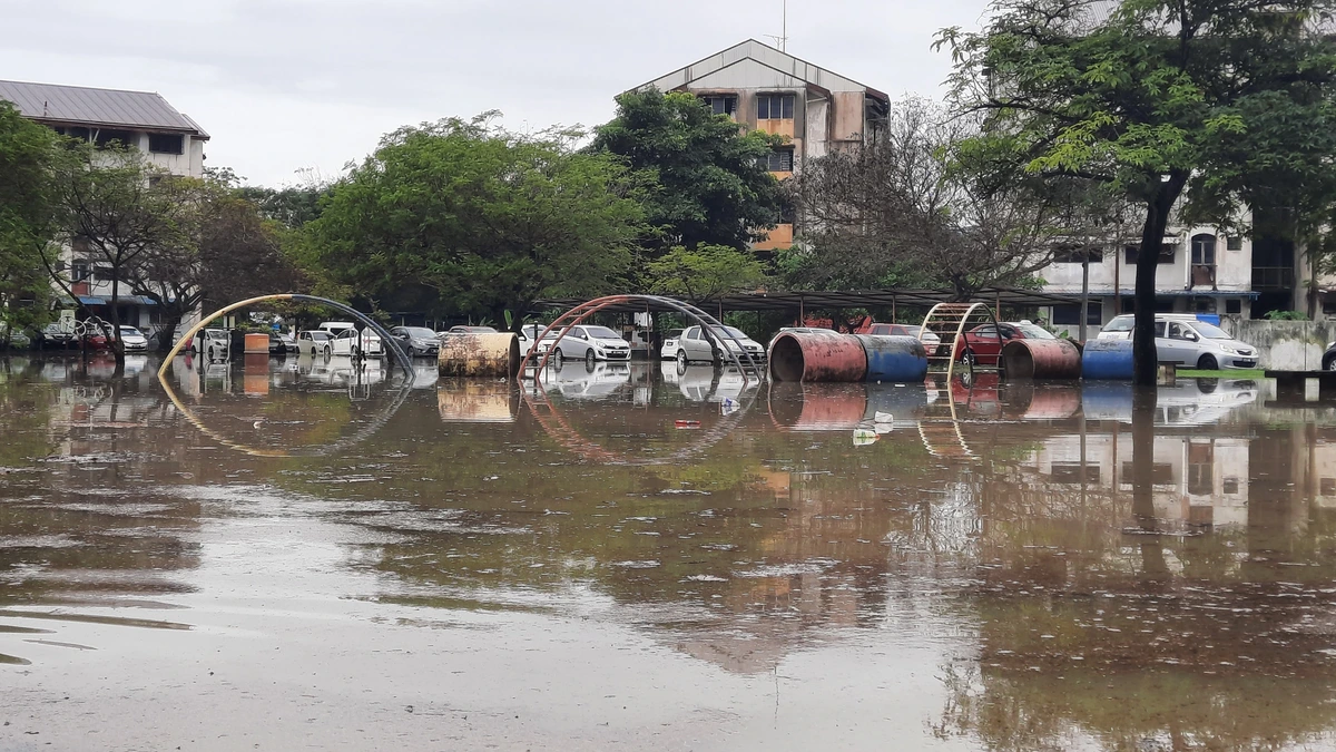 A flooded playground in Section 24, Shah Alam, with metal climbing arches and painted concrete pipes standing in brown floodwater, parked cars and a low-rise apartment block reflected behind