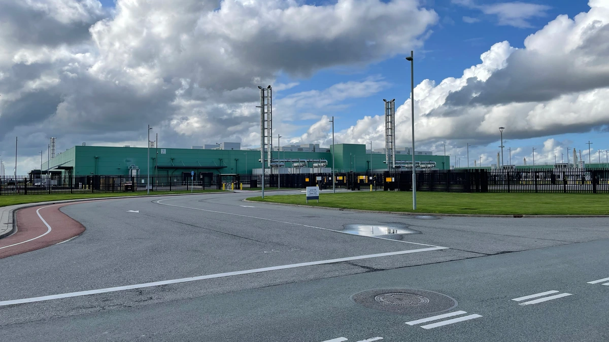 Green industrial data centre building behind a fence and grass verge under a blue sky with large clouds, seen from a road junction