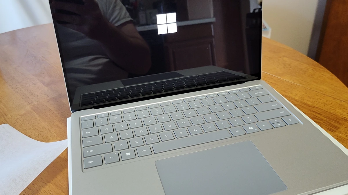 A silver Microsoft Surface Laptop open on a wooden table, showing the white Windows logo on its dark screen above a grey keyboard