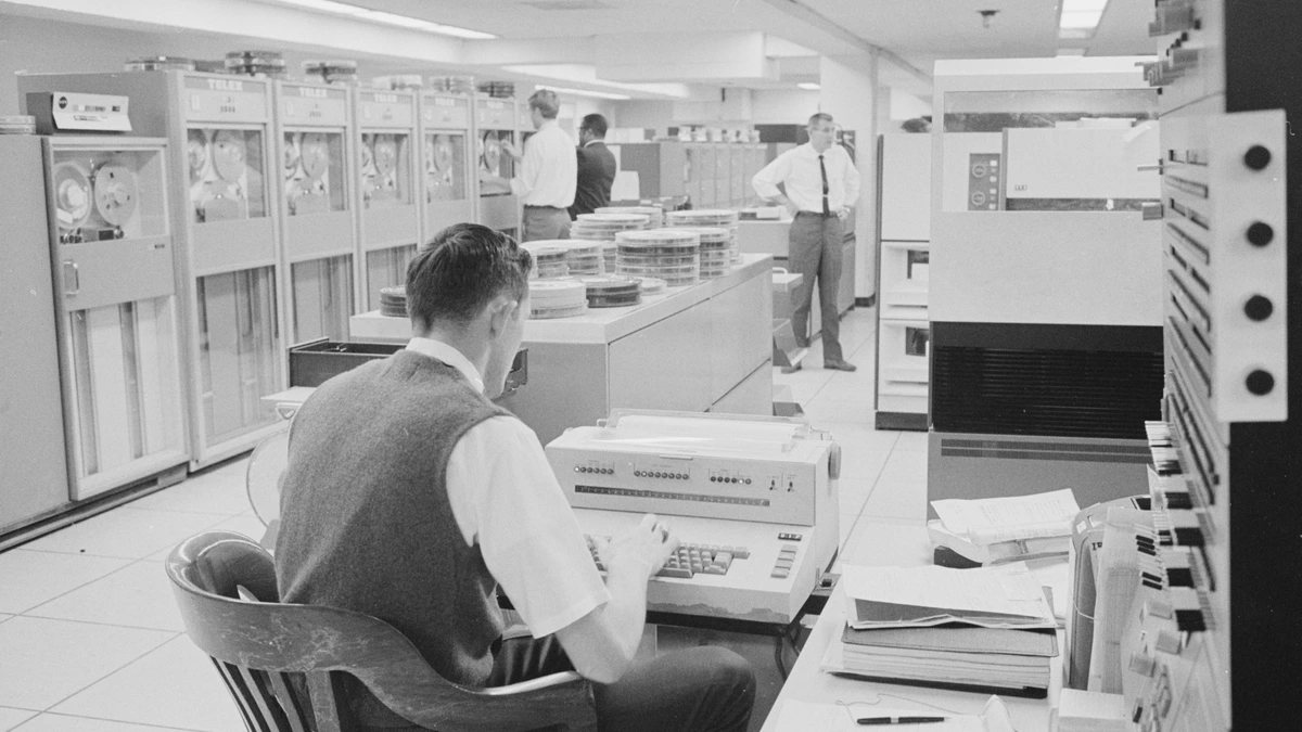 A 1960s computer room in which a man in a knitted vest types at a console printer, with banks of reel-to-reel tape drives along the left wall, stacks of tape reels on a long counter and three other staff standing among the cabinets