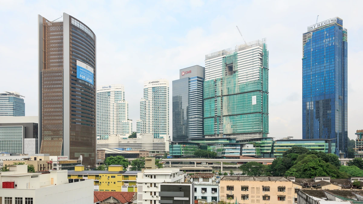A Kuala Lumpur skyline of office and hotel towers with a green-netted building under construction in the middle distance and older low-rise shophouses and homes in the foreground