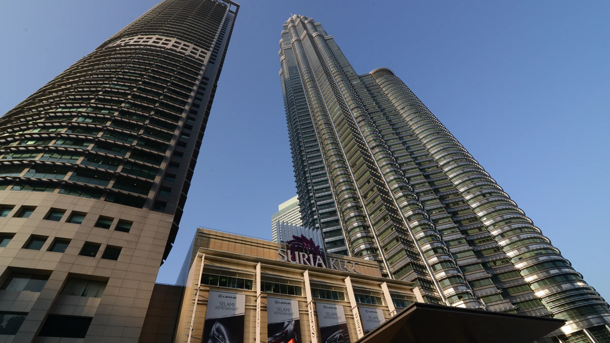The Petronas Twin Towers seen from street level beside a neighbouring office tower, their ribbed steel facades rising into a clear blue sky above the entrance canopy of the Suria KLCC mall
