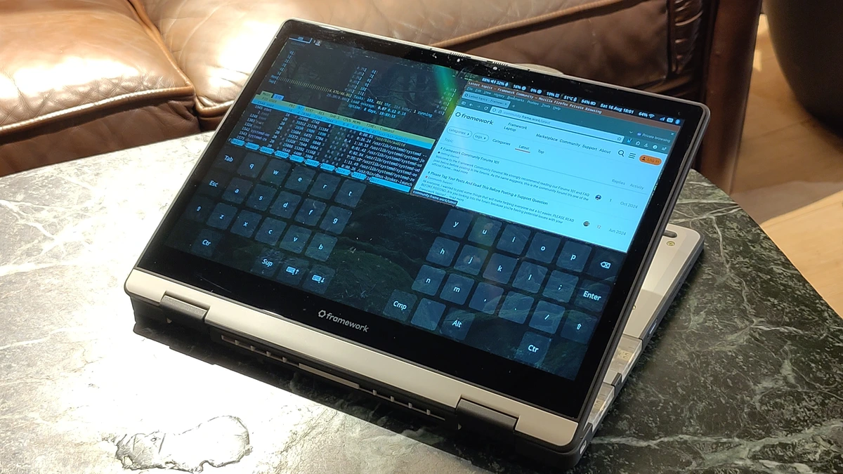 A Framework laptop folded into tablet mode on a green marble table in front of a brown leather sofa, its screen showing a Firefox browser window, a system monitor and an on-screen keyboard