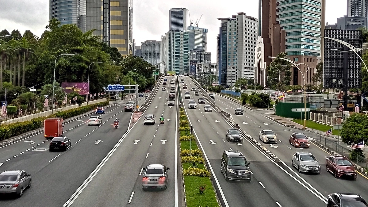Traffic on Jalan Tun Razak in Kuala Lumpur seen from above, with cars and motorcycles on a wide divided road lined with trees, Malaysian flags and office towers