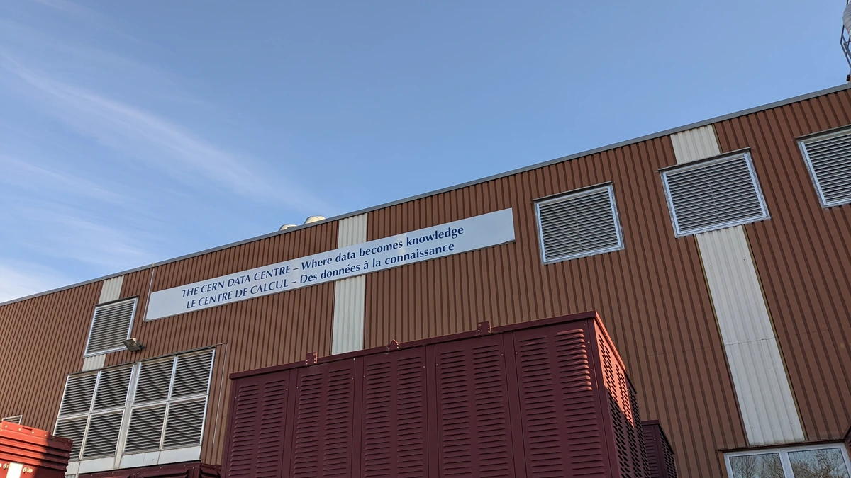 The corrugated exterior of the CERN data centre under a clear blue sky, with a long sign across the wall reading The CERN Data Centre, and dark red power equipment cabinets lined up in front of the building