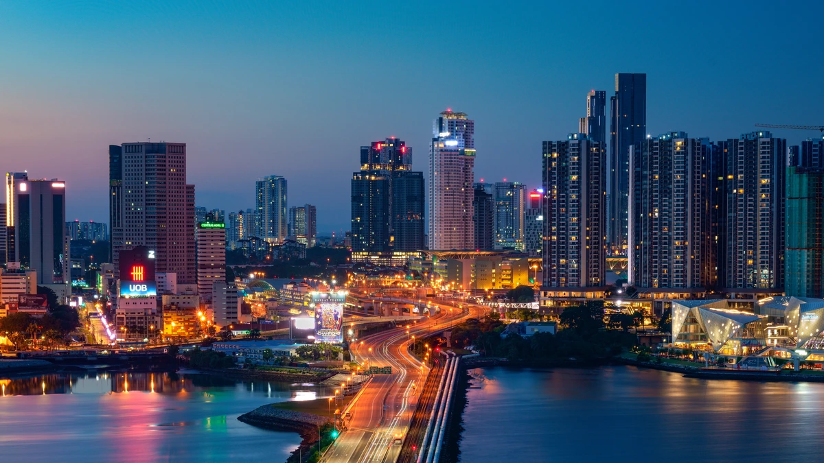 The Johor Bahru waterfront at dusk, with lit high-rise towers and construction cranes along the shoreline and light trails from traffic on the causeway below
