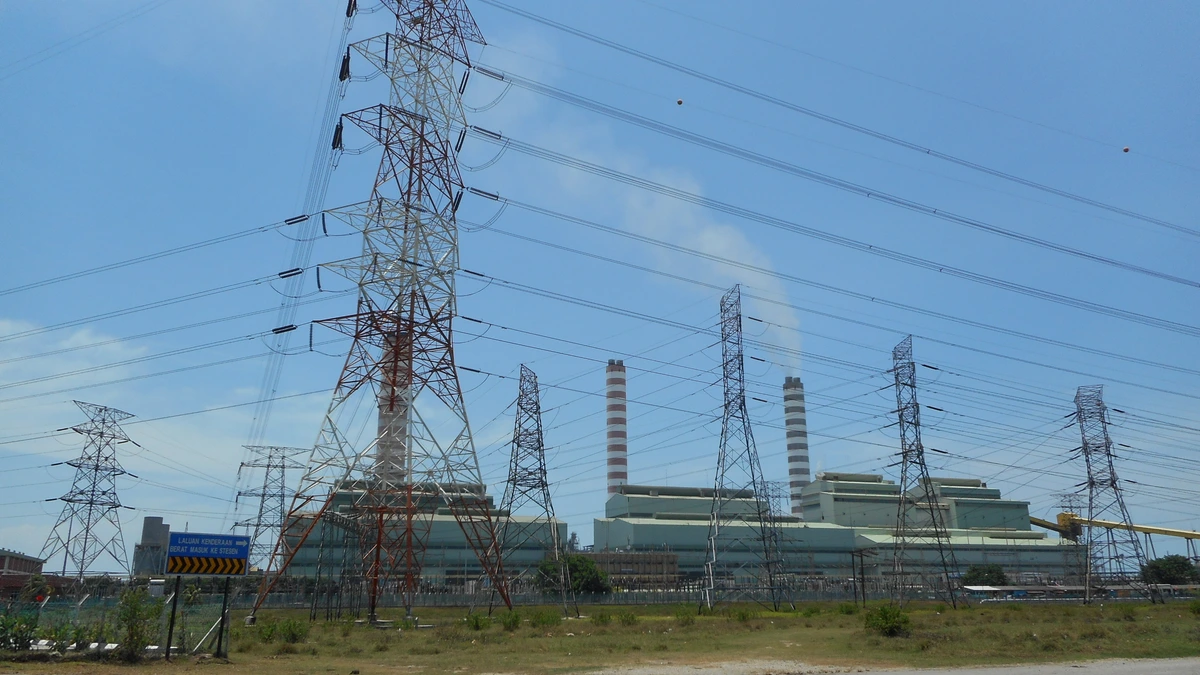 Transmission towers and overhead lines in the foreground with the chimneys and turbine halls of the Sultan Salahuddin Abdul Aziz power station behind them under a bright sky