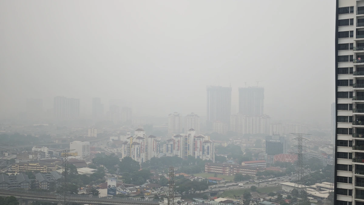 Kuala Lumpur apartment towers, cranes and roads fading into thick grey haze under a white sky, with a high-rise balcony edge on the right