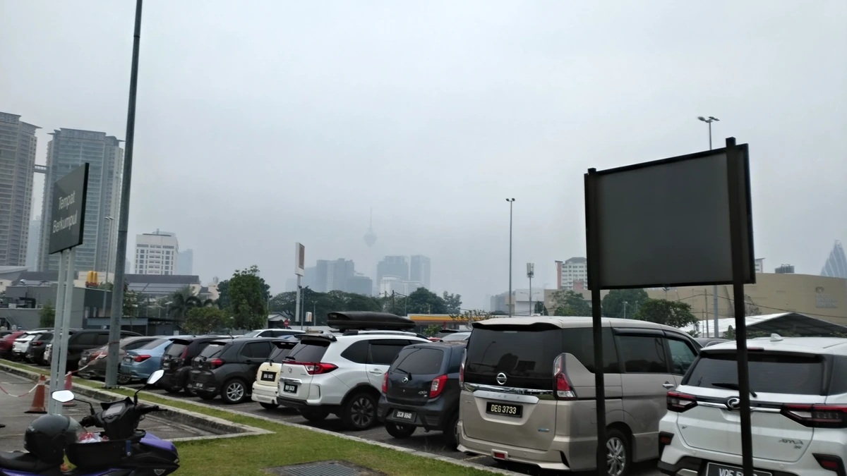 Cars parked in rows on a hazy morning with a motorcycle on the verge, grey sky, and high-rise buildings and a thin tower faint in the haze behind