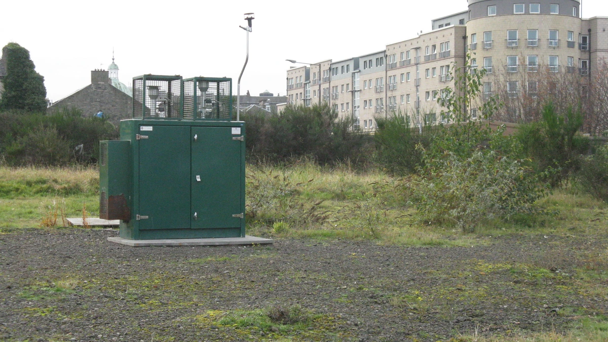 A green metal air quality monitoring cabinet with caged sampling inlets on its roof and a thin mast, standing on gravel in rough grassland beside modern flats