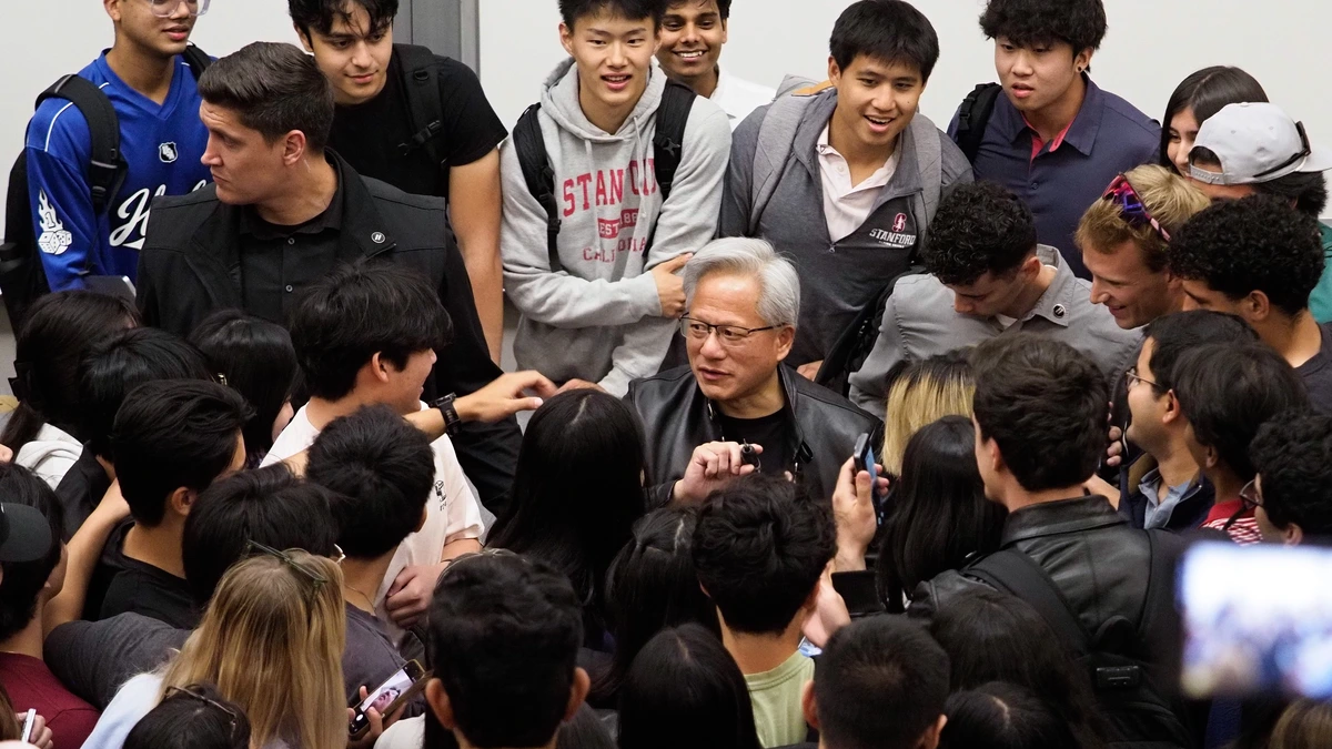 A grey-haired man with glasses in a black leather jacket is surrounded by a crowd of students, some holding up phones, in a lecture hall