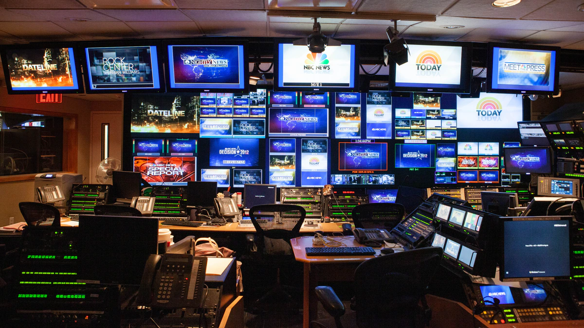 A television network control room with a wall of dozens of monitors showing programme logos and live feeds above two long desks of mixing panels, keyboards and telephone handsets