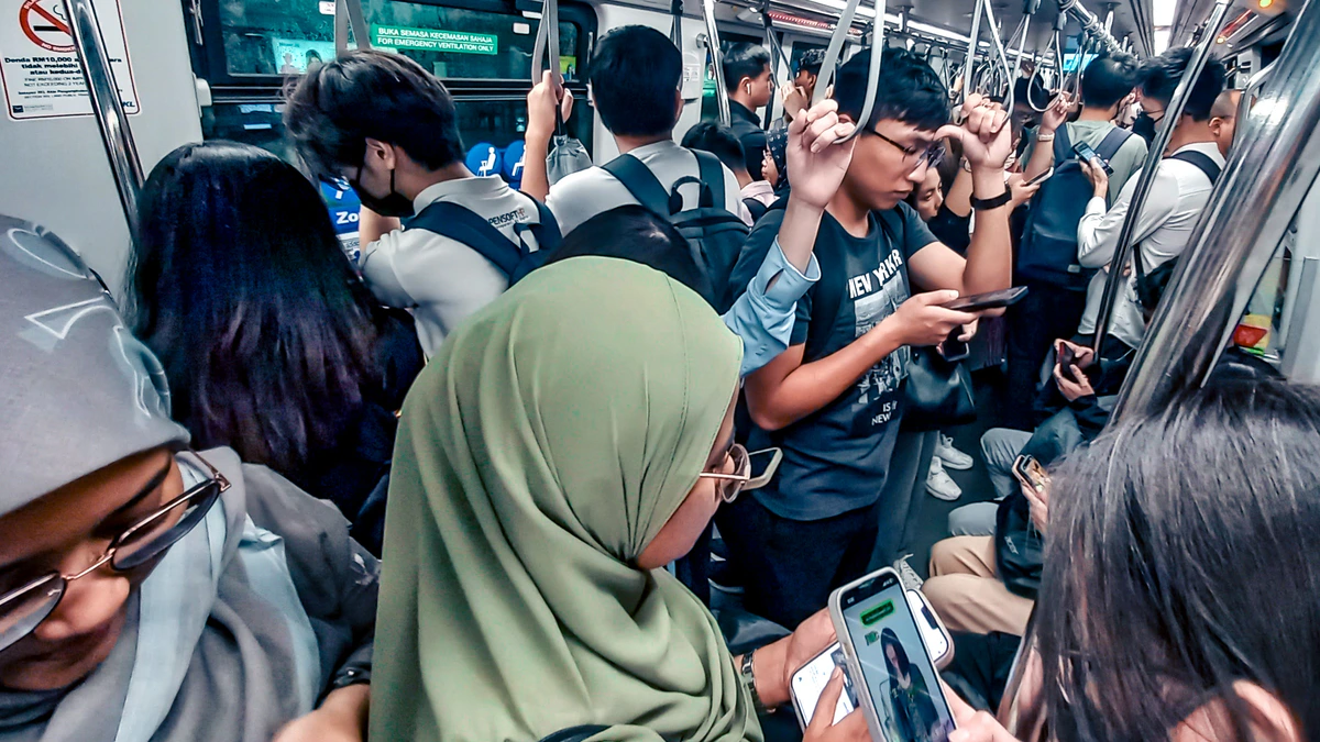 A crowded Kuala Lumpur train carriage of standing passengers holding overhead rails, several looking down at phones, with one screen in the foreground showing a video of a person
