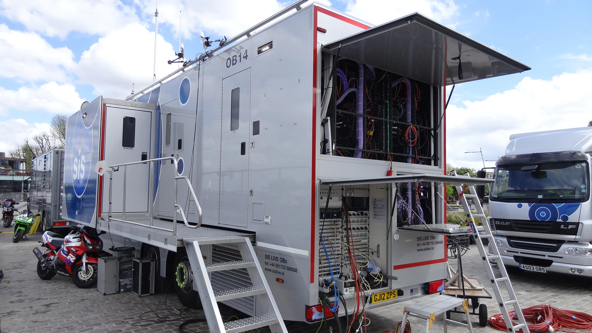 A silver outside broadcast truck parked on cobbles with its side panel raised, exposing racks of cabling and connectors, an access ladder and coiled cables on the ground beside it