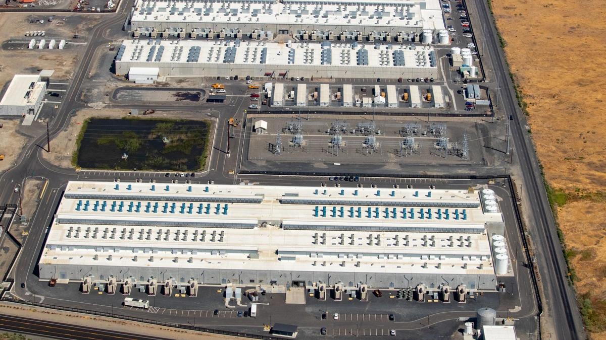 Aerial view of three large flat-roofed Amazon Web Services datacentre buildings with rows of cooling units, a fenced electrical substation and a car park, next to a highway and dry grassland in Oregon