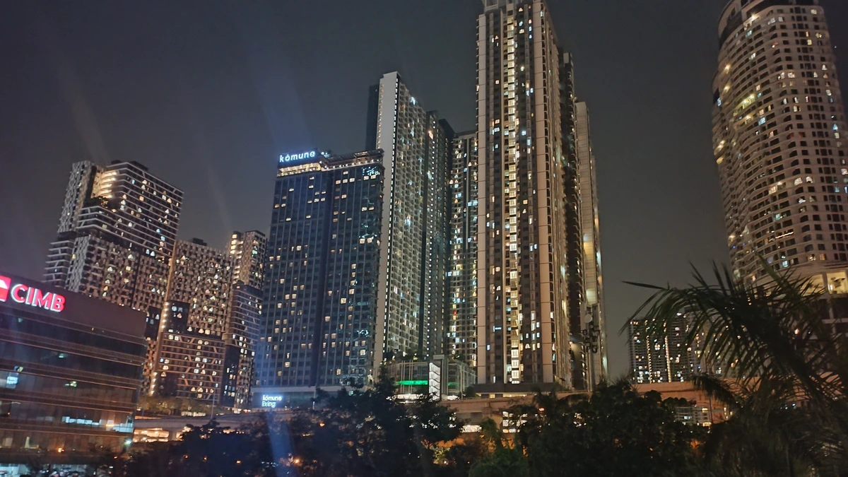 Lit office and residential towers of Bangsar South in Kuala Lumpur at night, with a bank sign on a low building on the left and dark trees in the foreground