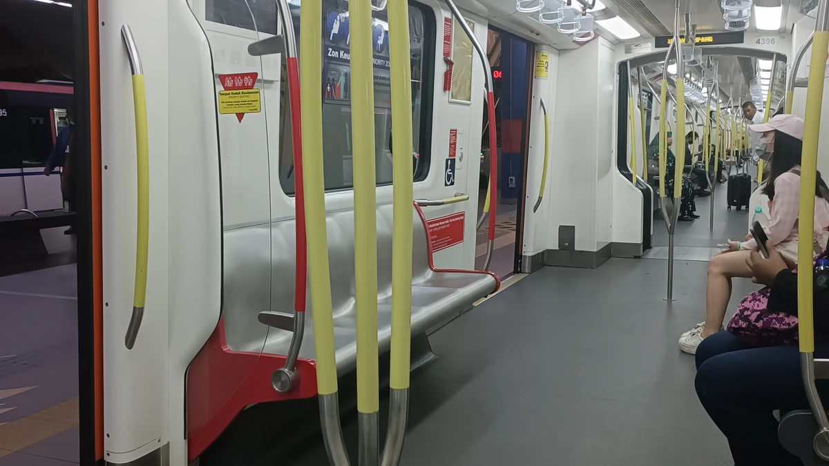 Inside a Sri Petaling Line LRT carriage in Kuala Lumpur, with yellow grab poles in the foreground, an open door at left and a few seated passengers at right, one holding a phone