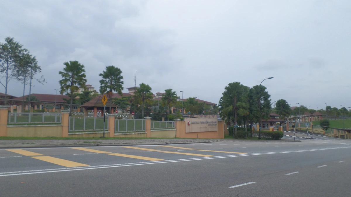 The fenced entrance and signboard of Hospital Permai in Johor Bahru, a low-rise hospital complex with red-tiled roofs and palm trees, seen across an empty road under a grey sky