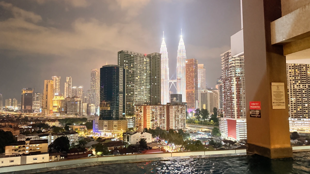 Kuala Lumpur skyline at night with the lit Petronas Twin Towers rising above apartment blocks, seen across the edge of a rooftop pool