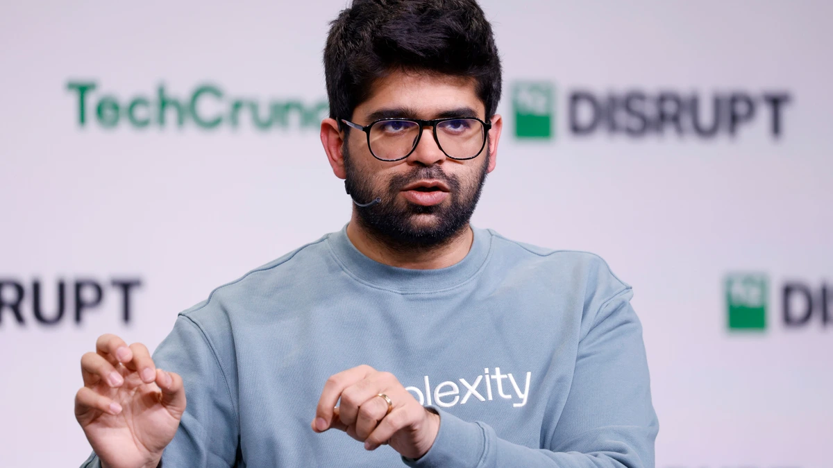 Perplexity chief executive Aravind Srinivas in glasses and a grey Perplexity sweatshirt, gesturing as he speaks on stage in front of a TechCrunch Disrupt backdrop