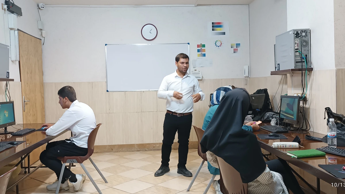 An instructor in a white shirt standing and speaking in a computer training room while seated learners work at desktop monitors along the wall
