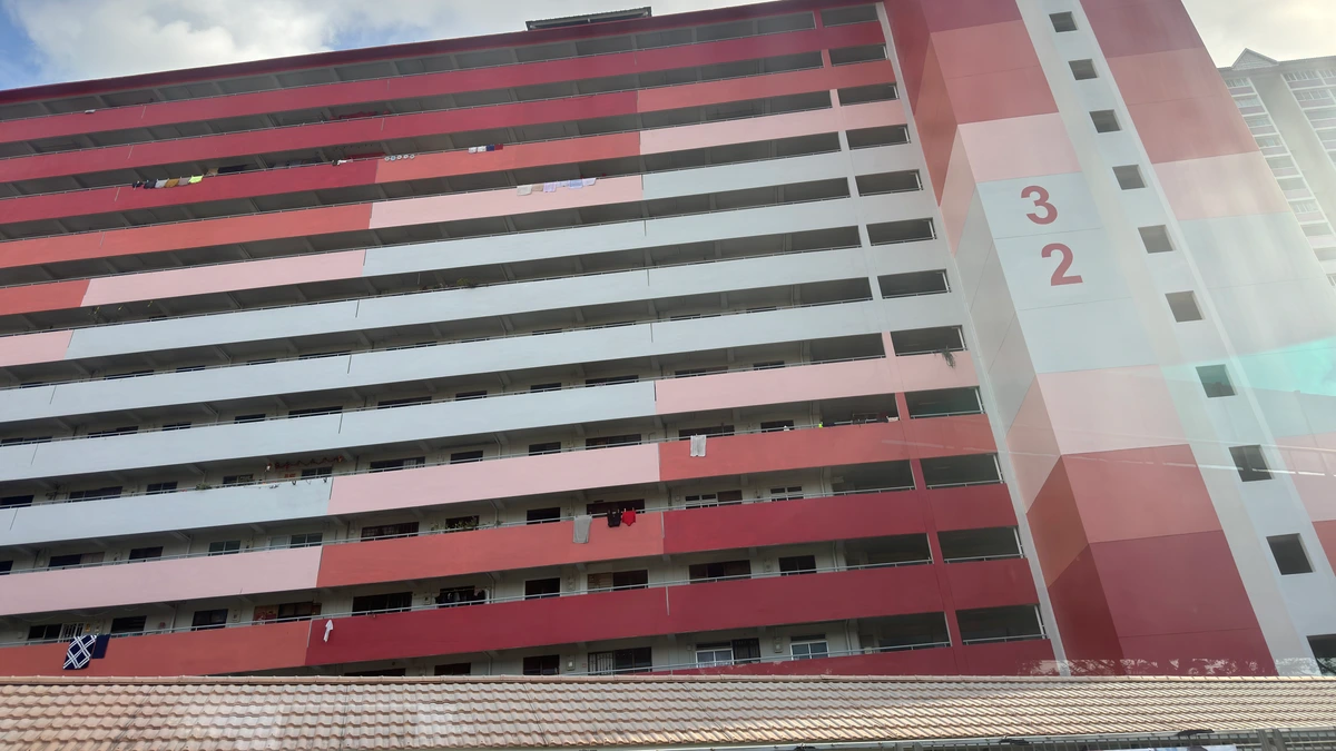 A tall public housing block in Singapore painted in bands of red, pink and off-white, its open corridors and laundry poles visible, seen from street level beneath a tiled shelter roof