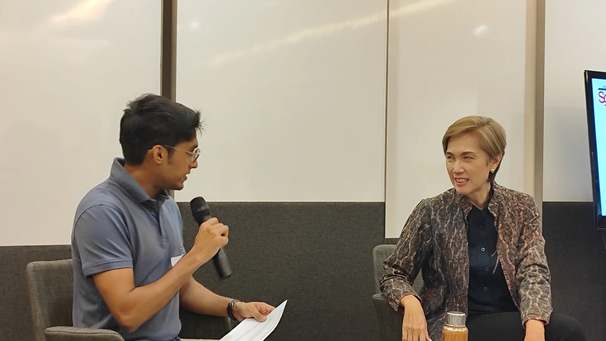Josephine Teo in a patterned jacket sitting on a low armchair in conversation with a younger man in a blue polo shirt who holds a microphone and a sheet of notes
