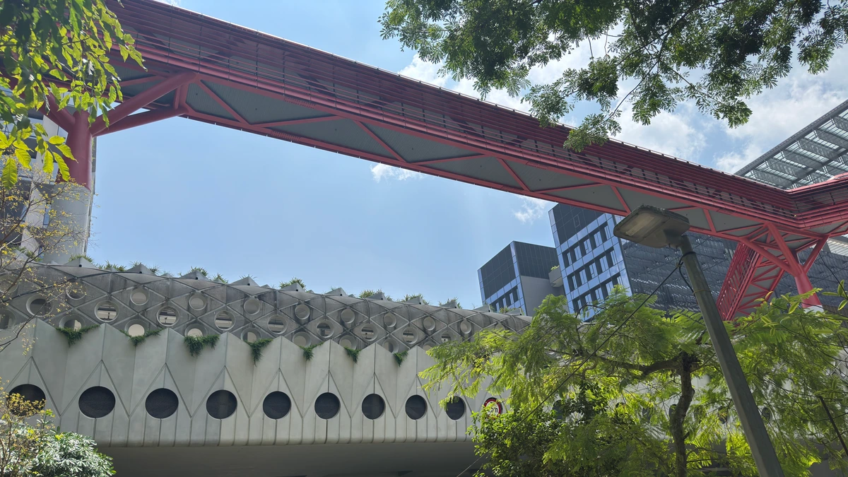 A red steel pedestrian bridge crossing above a pale concrete building with a honeycomb facade of circular openings planted with greenery, with taller blocks and trees behind it