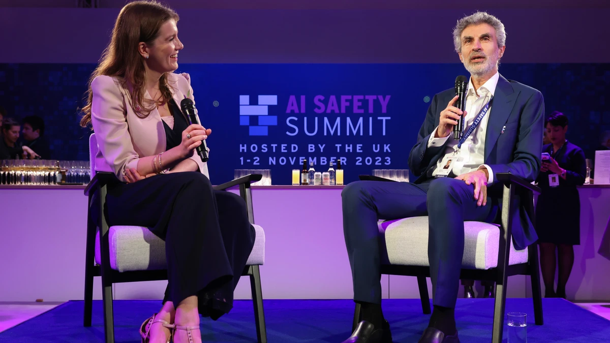 Yoshua Bengio, seated on stage and holding a microphone, speaks beside then UK Science Secretary Michelle Donelan in front of a backdrop reading AI Safety Summit, Hosted by the UK, 1-2 November 2023