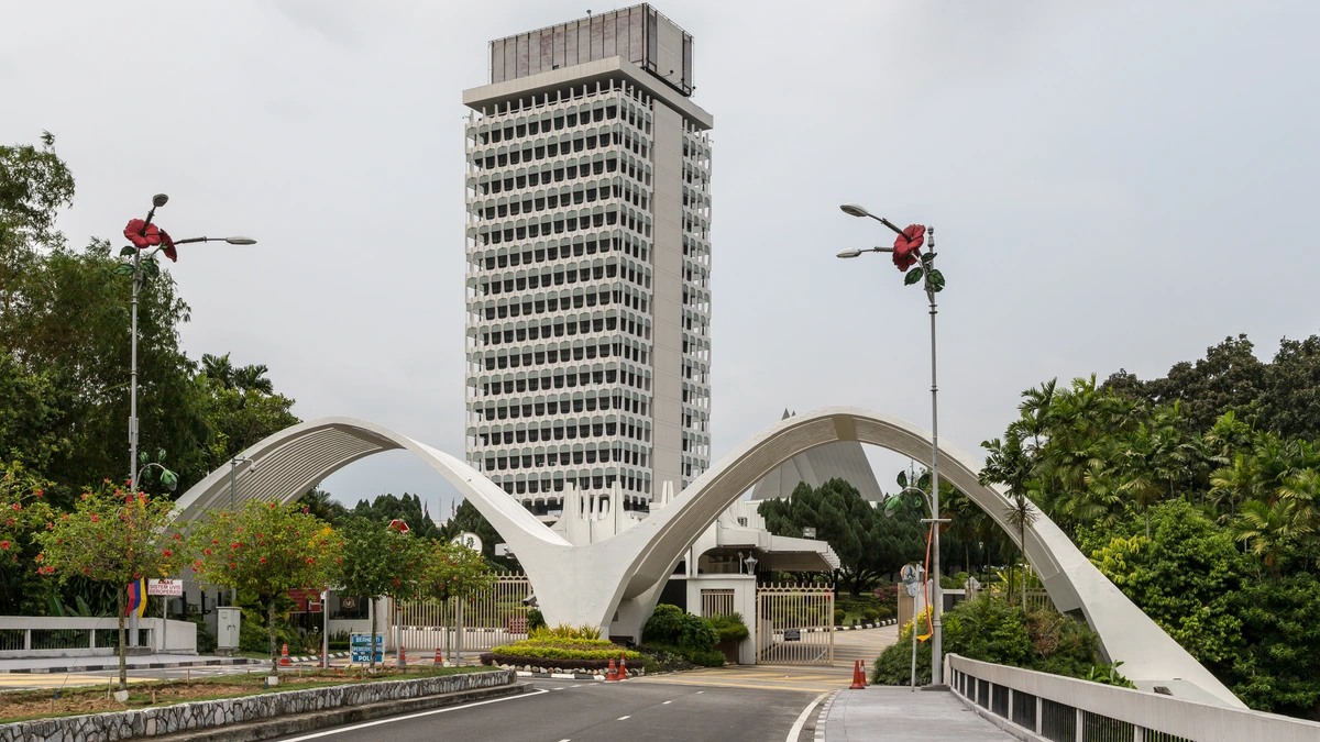 The white tower block of Malaysia's Parliament in Kuala Lumpur, seen down the entrance road through a white twin-arch gateway, with hibiscus-shaped street lamps and tropical trees on either side