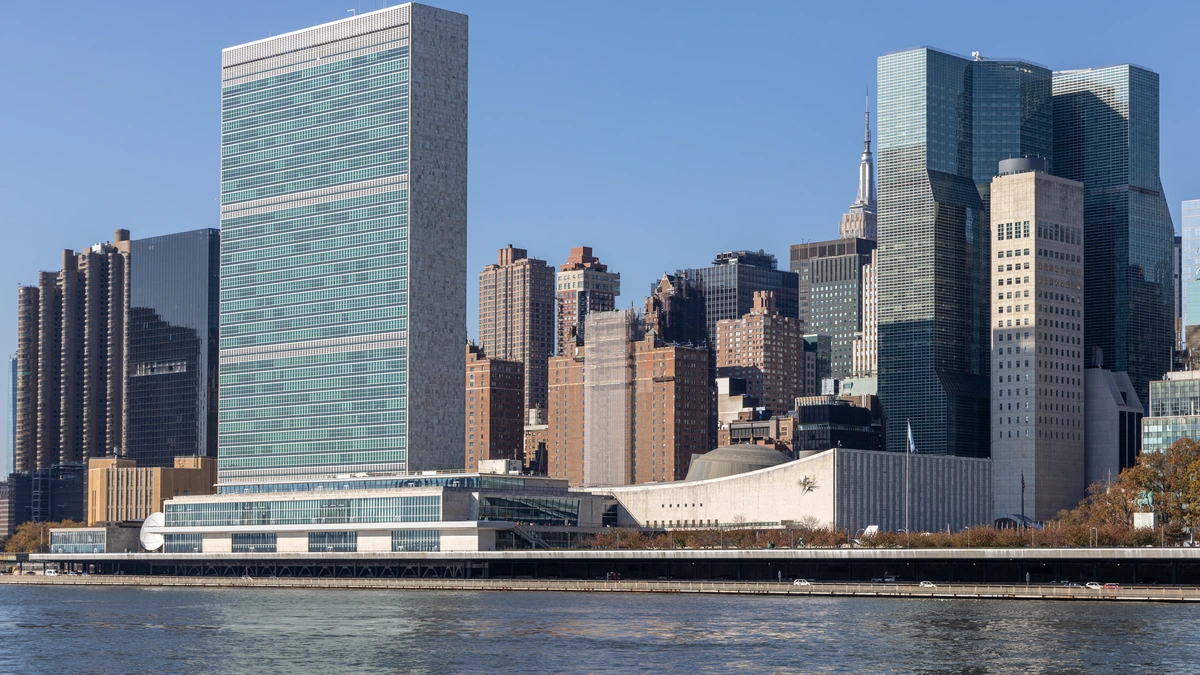 The tall glass UN Secretariat tower and the low, curved General Assembly building on the East River shore in Manhattan under a clear blue sky, with the Empire State Building's spire behind