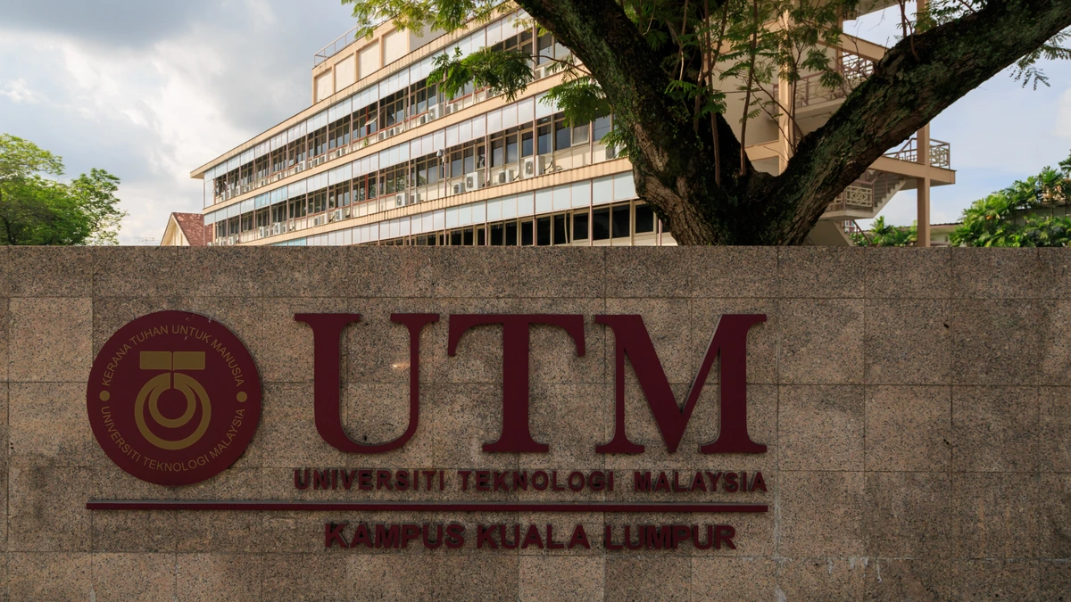 A maroon UTM sign on a stone wall reading Universiti Teknologi Malaysia, Kampus Kuala Lumpur, with a campus building behind it