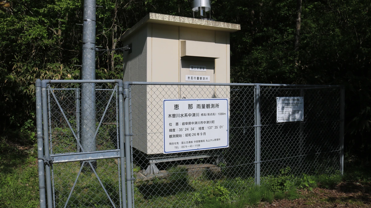 A small beige rain gauge station behind a chain-link fence, with a white information sign in Japanese