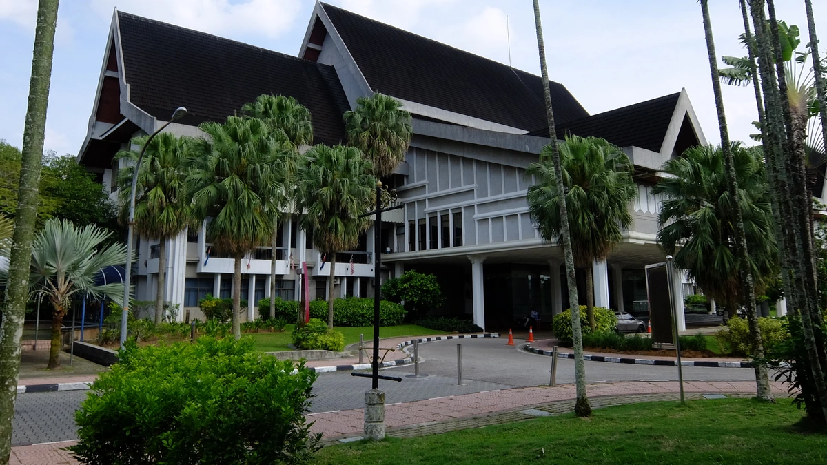 The Sultan Ibrahim Chancellor Building at UTM Skudai, a white building with a dark pitched roof and wide eaves, framed by tall palm trees and a paved path