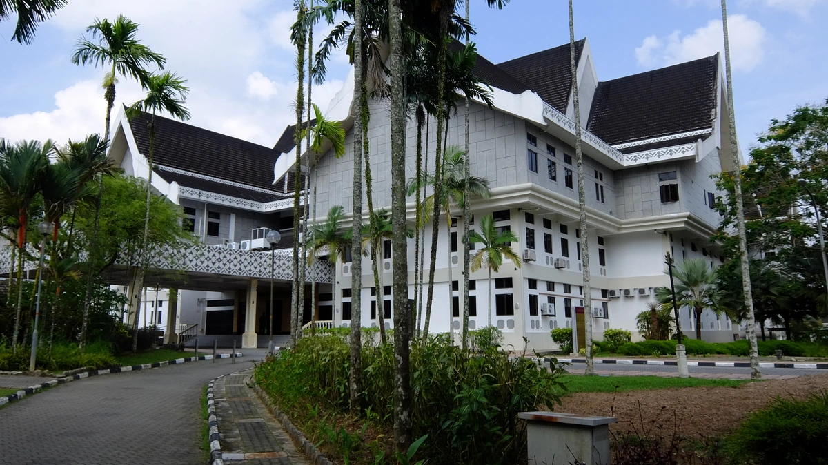 Sultan Iskandar Hall at UTM Skudai, a white multi-storey building with dark roofs rising behind tall palm trees under a blue sky with clouds
