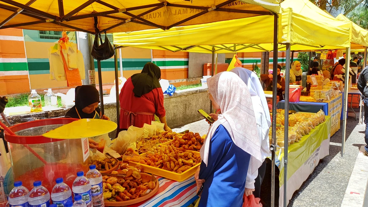 A woman in a pink headscarf looks at her phone while standing at a street food stall under yellow canopies at a market on Jalan Benteng, Kuala Lumpur, with trays of fried snacks and bottled water on the counter